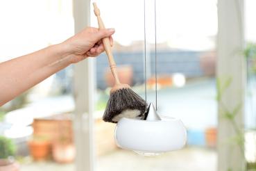 Preview: Ein langer Staubpinsel mit grau-hellem Besatz befreit eine weiße Deckenlampe von Staub.