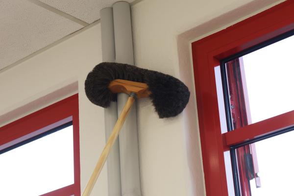 Der Wiener Wandbesen mit schwarzen Besatz befreit eine weiße Wand in der Nähe eines grauen Heizungsrohrs und roten Fensterrahmen vom Staub.
